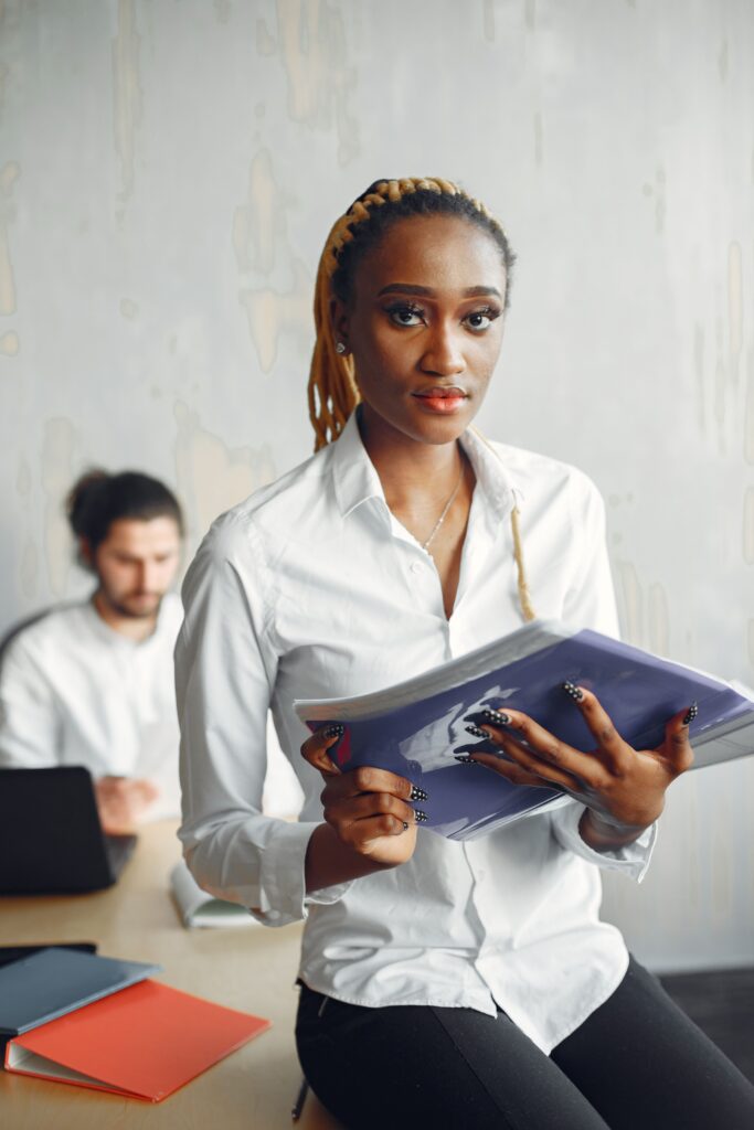 Young professional woman holding a folder in a modern office environment with a colleague working in the background.